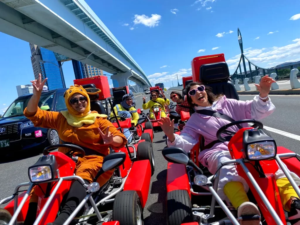 Group of people in red go-karts riding on a city road, some in colorful costumes and sunglasses, waving at the camera under a large bridge.