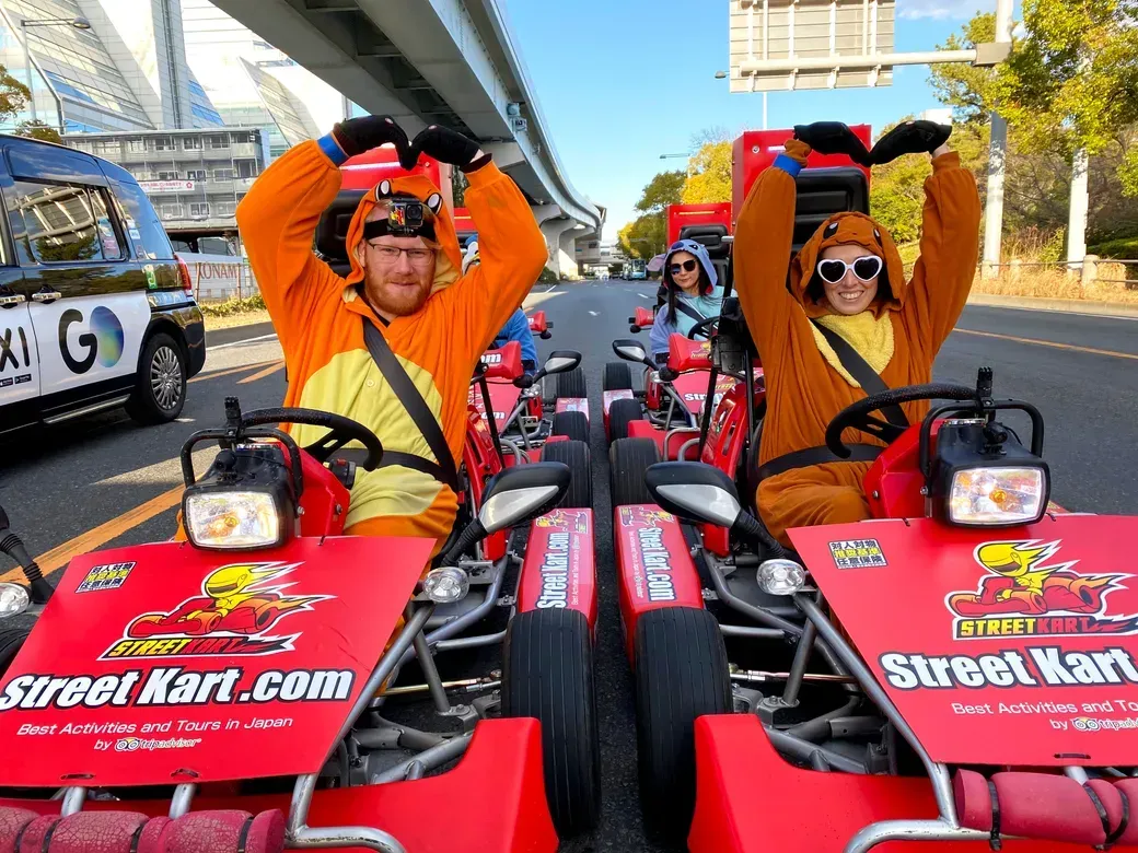People in orange animal onesies pose in go-karts on a city street, forming a heart shape with their arms above the steering wheels, Street Kart branding visible.